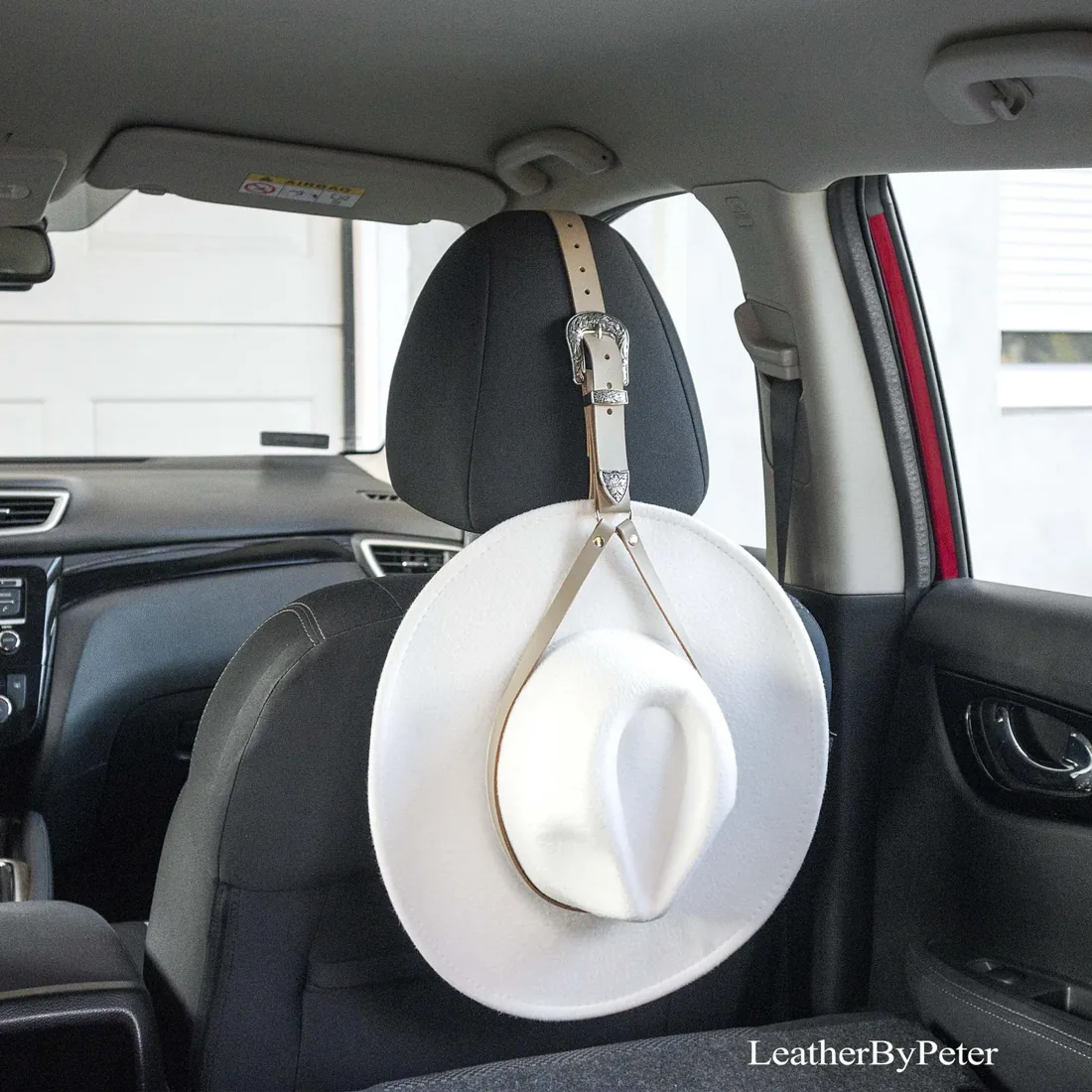 Gray leather cowboy hat holder with decorative silver buckle, attached to a car headrest and holding a white cowboy hat.