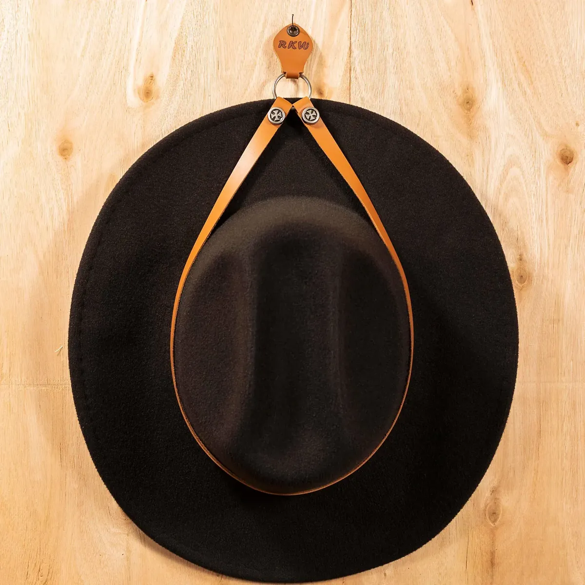 Personalized tan leather cowboy hat holder with metal eyelet and decorative star rivets, holding a dark cowboy hat on a wooden wall