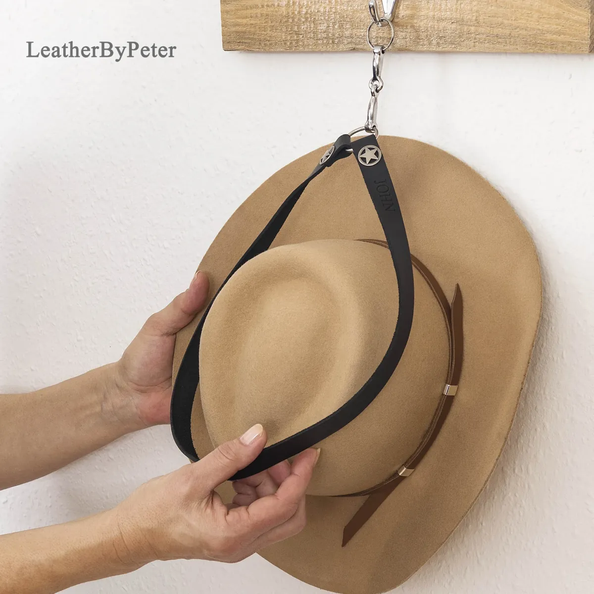 Black leather cowboy hat holder with a miniature snaffle bit decorative fitting, hanging from a hook with a tan cowboy hat