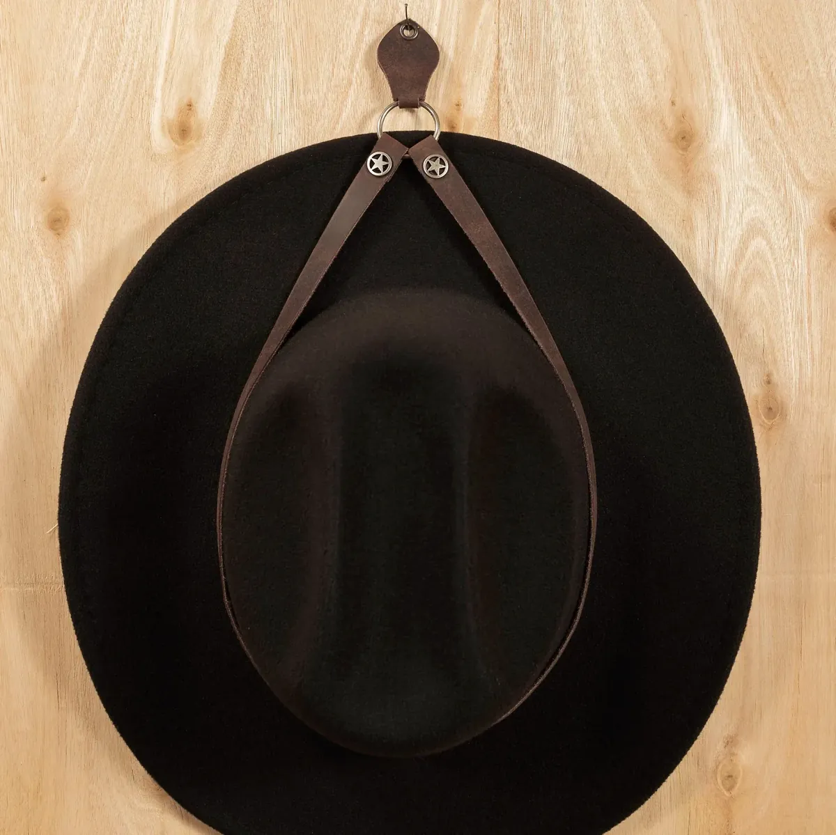 Dark brown leather cowboy hat wall holder with eyelet, metal ring and western star details on a wooden wall